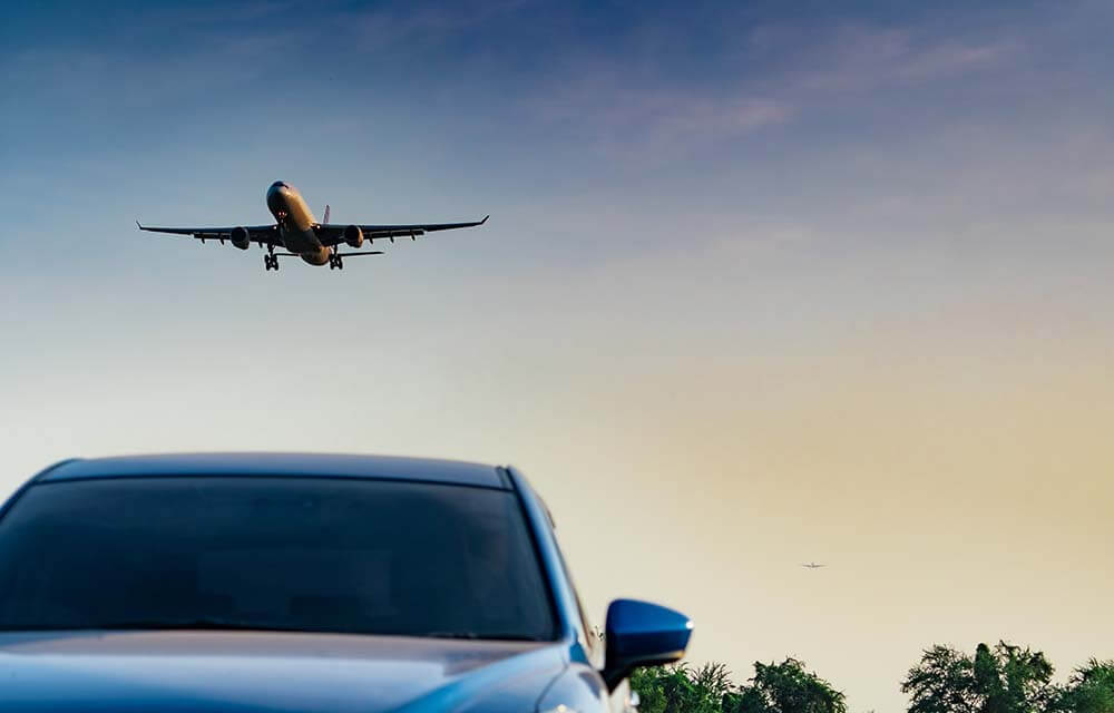 Commercial airplane flying above luxury car hood symbolizing air and car travel options with blurred city background