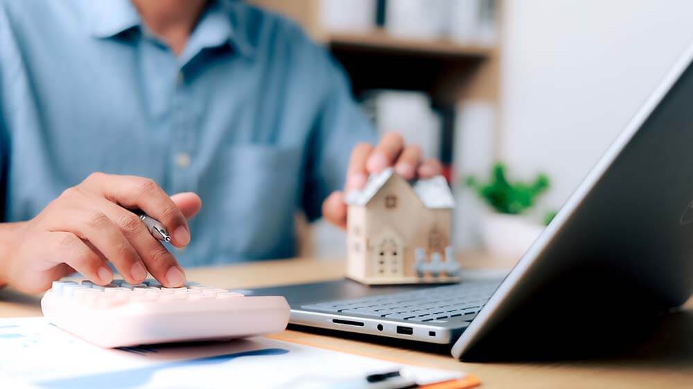 Man calculating property purchase costs on laptop with money and documents on table surface