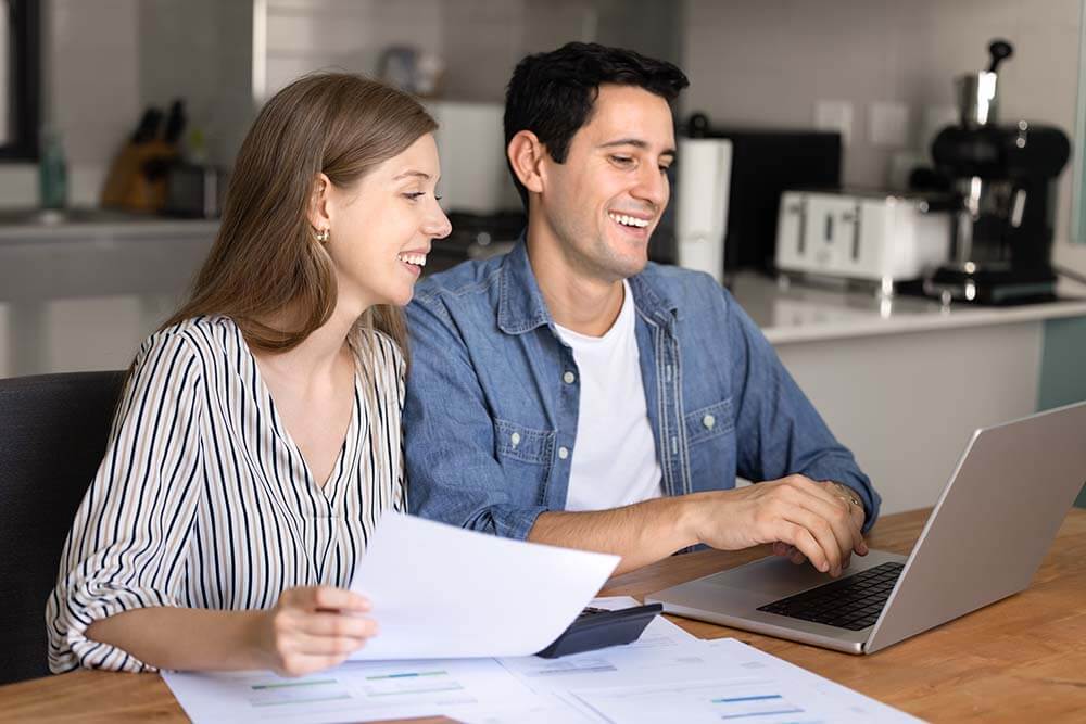 Happy couple sitting together at wooden table reviewing property transaction steps with laptop and documents around them