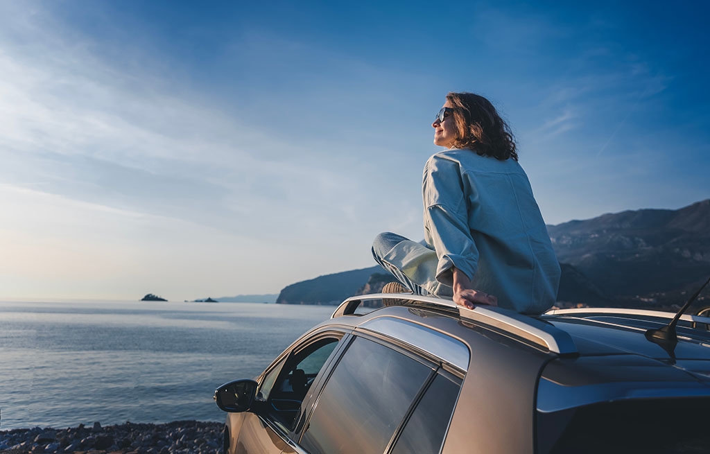 Woman sits on car roof, viewing sea landscape, enjoying the freedom of life in Bulgaria